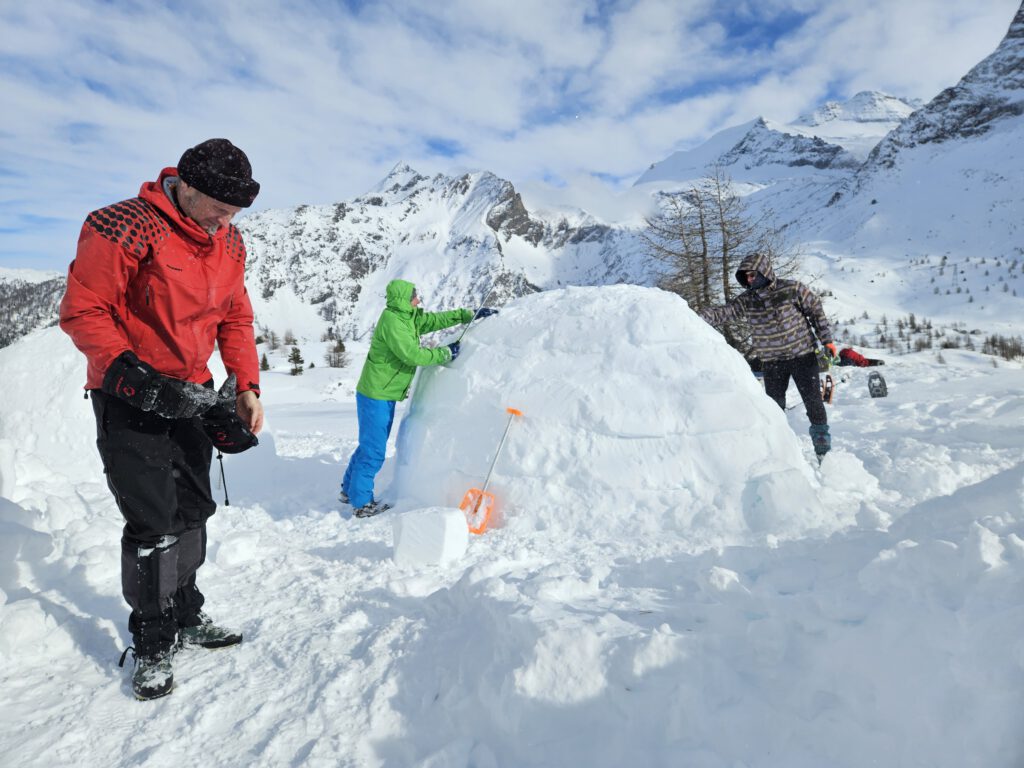 Drei Personen bauen ein Iglu in schneebedeckter Berglandschaft
